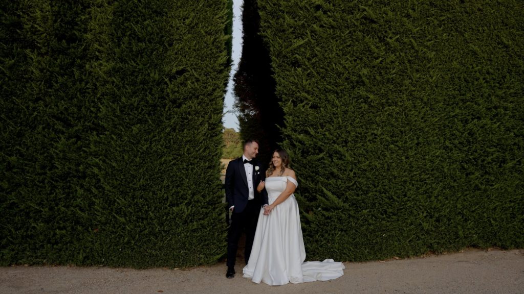Wedding photography of bride and groom standing between tall manicured hedges at a garden venue.